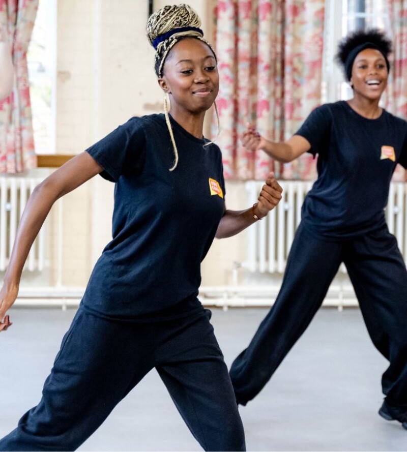 Two young performers in black Stagecoach T-shirts pose mid-dance in a studio, with one dancer in the foreground extending their arms and smiling.