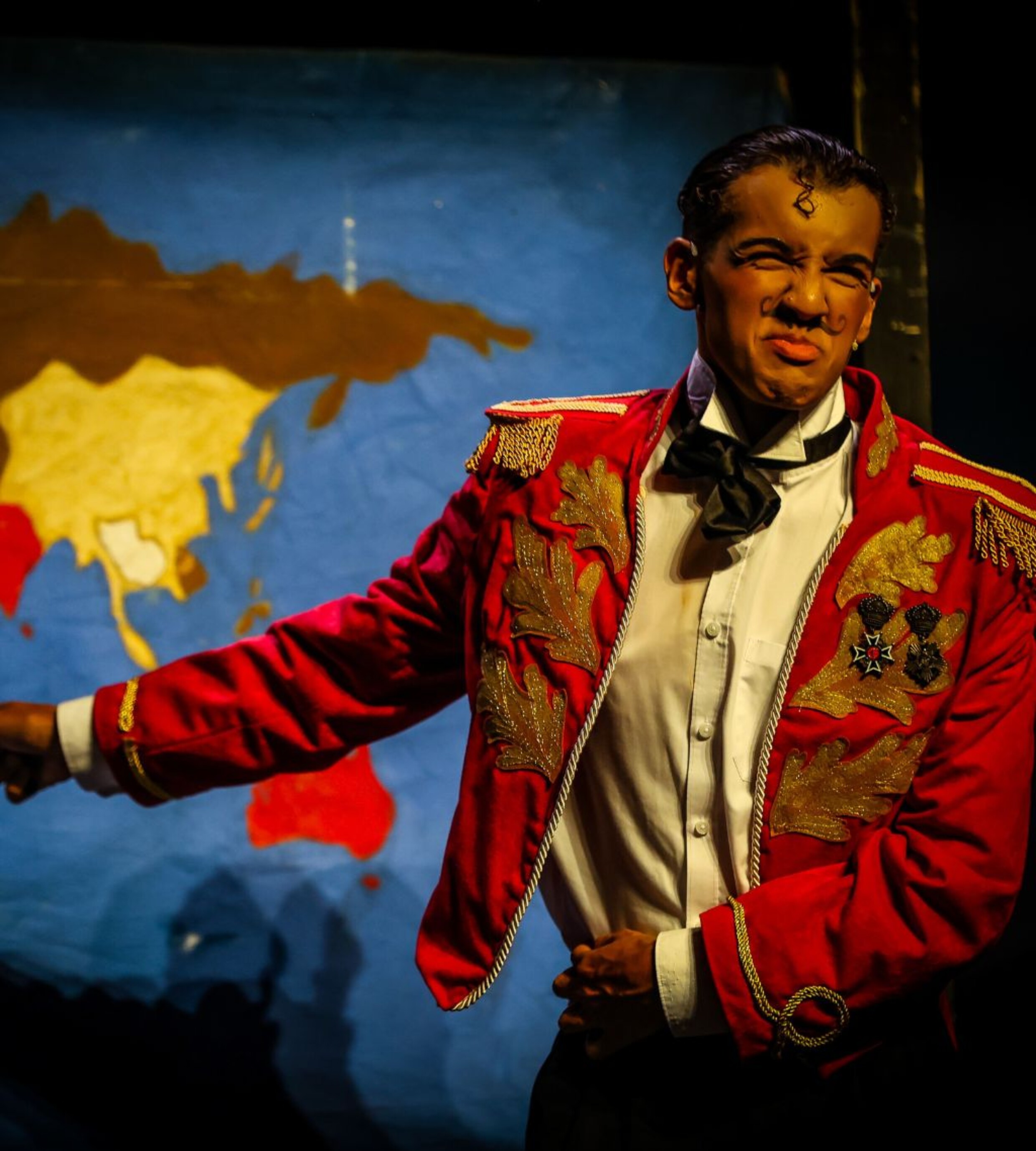 A young performer in an ornate red jacket and black bow tie acts expressively on stage in front of a painted map backdrop during the Chitty Chitty Bang Bang summer showcase.