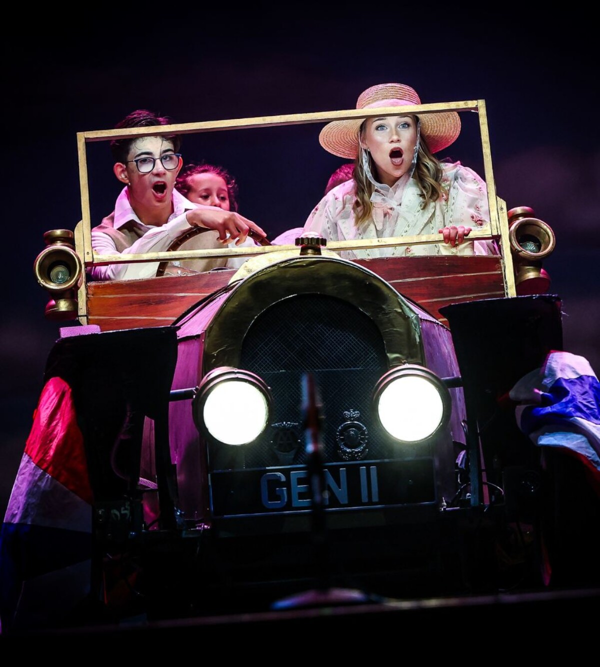 Performers sit inside a vintage car prop with bright headlights, looking surprised and excited under stage lighting.