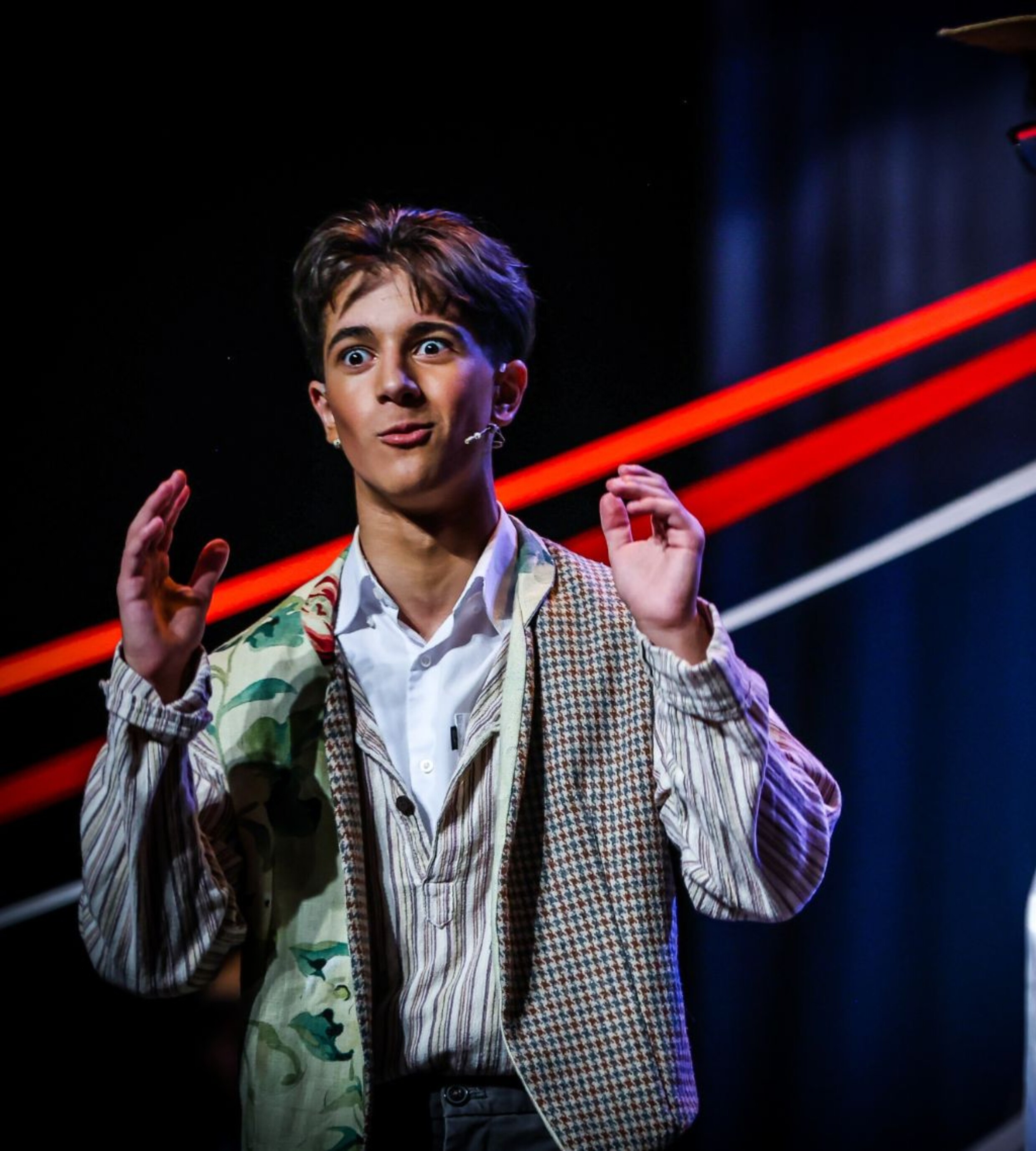 A young performer wearing a patterned waistcoat and headset microphone gestures animatedly on stage with bold red lighting behind them.