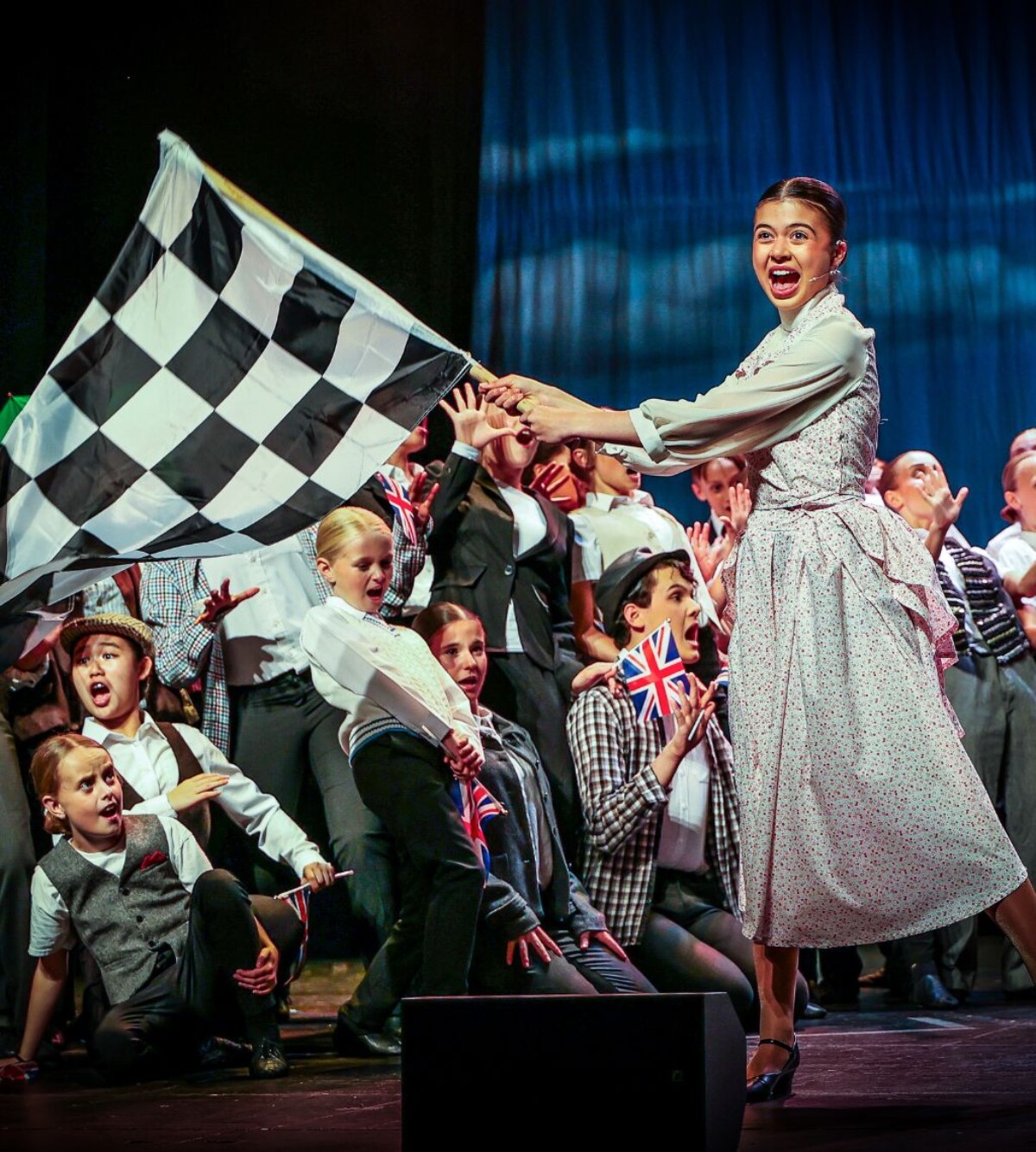 A young performer waves a large black-and-white chequered flag as the ensemble cheers behind her with Union Jack flags.