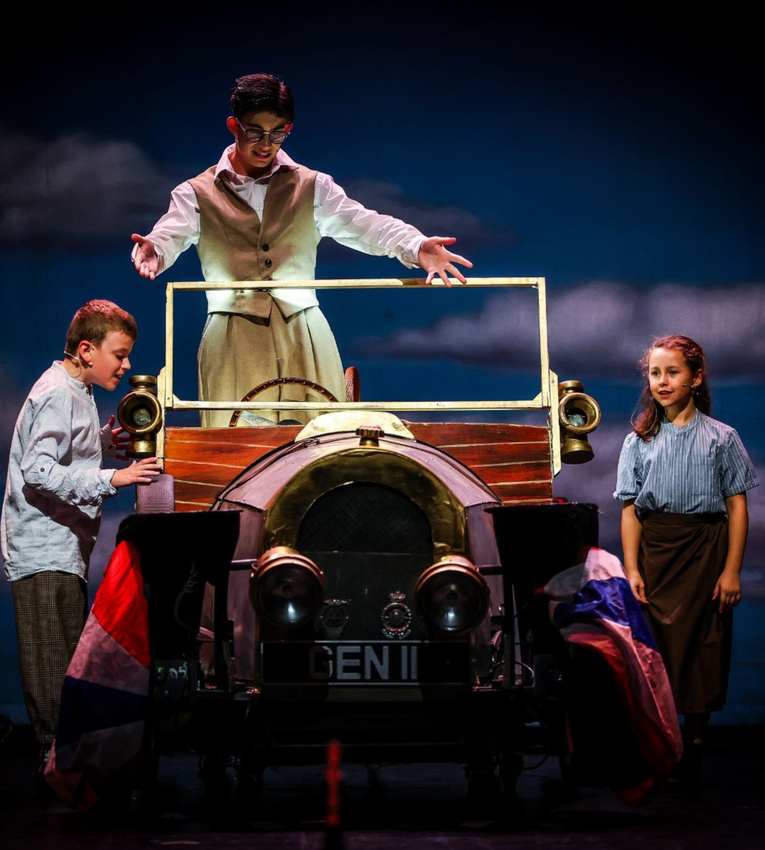 Three young performers gather around a vintage car prop on stage, with one standing inside the car and two beside it.