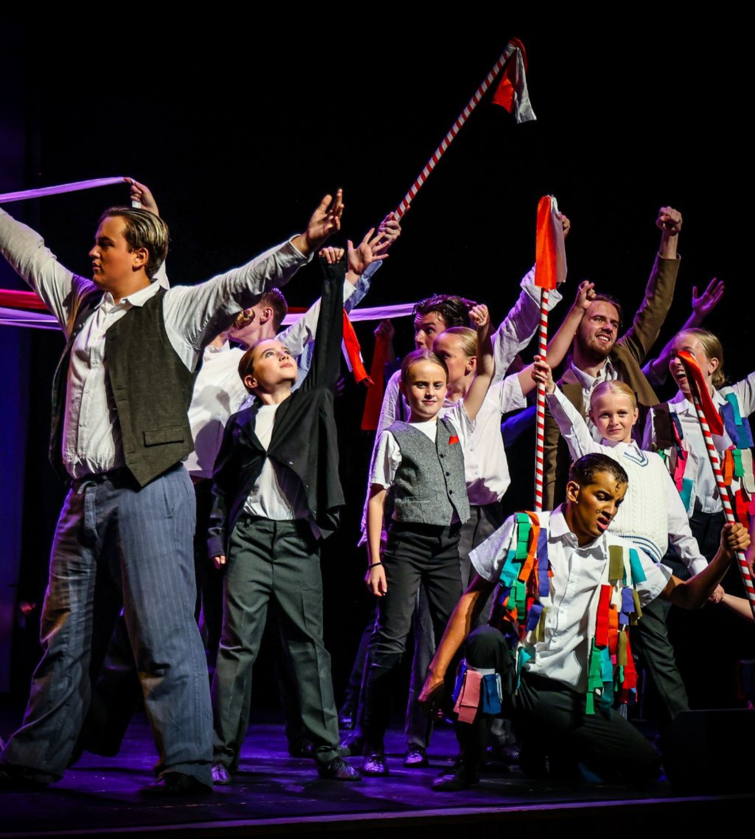 A large ensemble of performers pose energetically on stage, raising striped poles, ribbons and flags during a lively musical number.