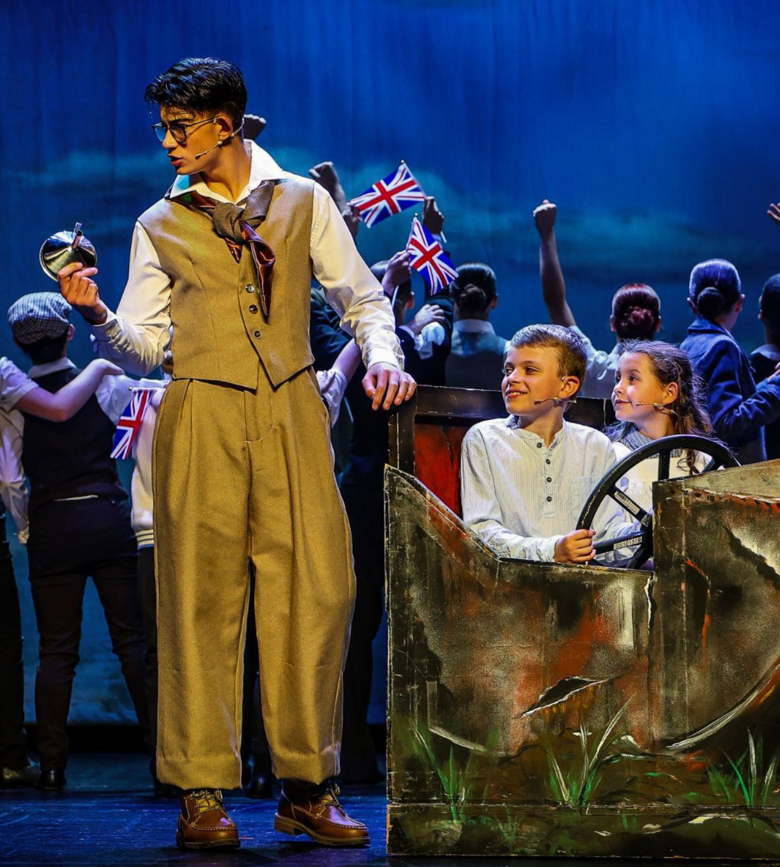 A young performer holding a pocket watch stands beside a rustic car prop, while two young performers sit inside and the ensemble waves Union Jack flags in the background.